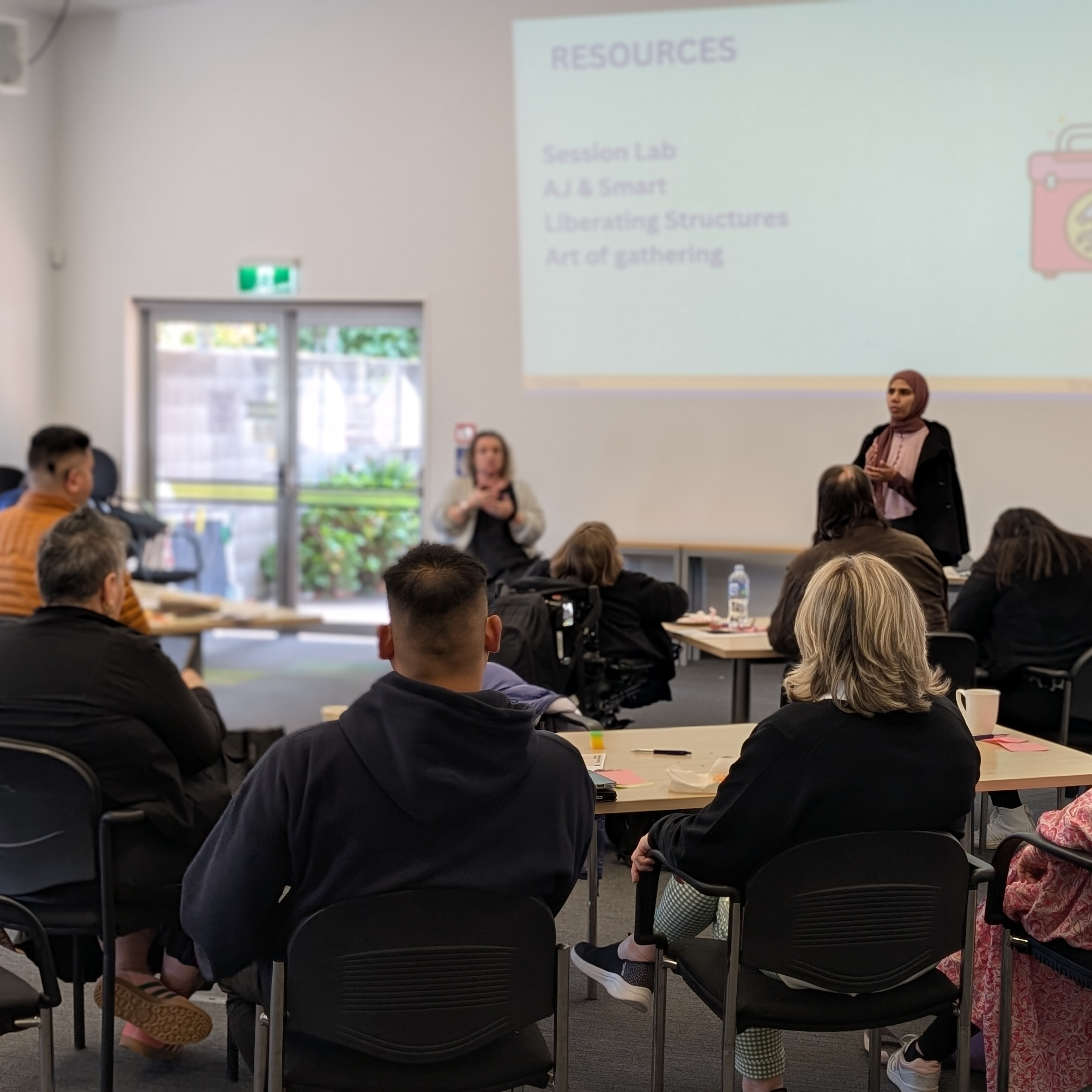 A woman presents to a seated group in a classroom setting, with a screen displaying a slide titled “RESOURCES” and a list of tools.